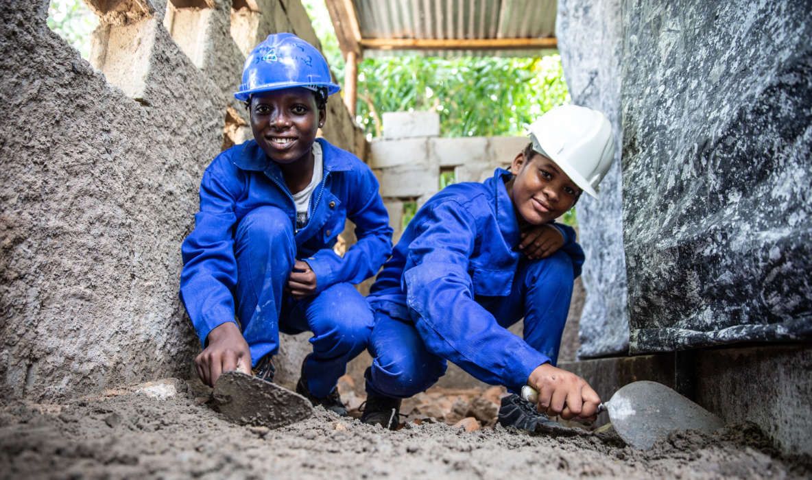 Two trainees in blue overalls and hard hats practising construction skills at ADPP Escola Vocacional Nacala